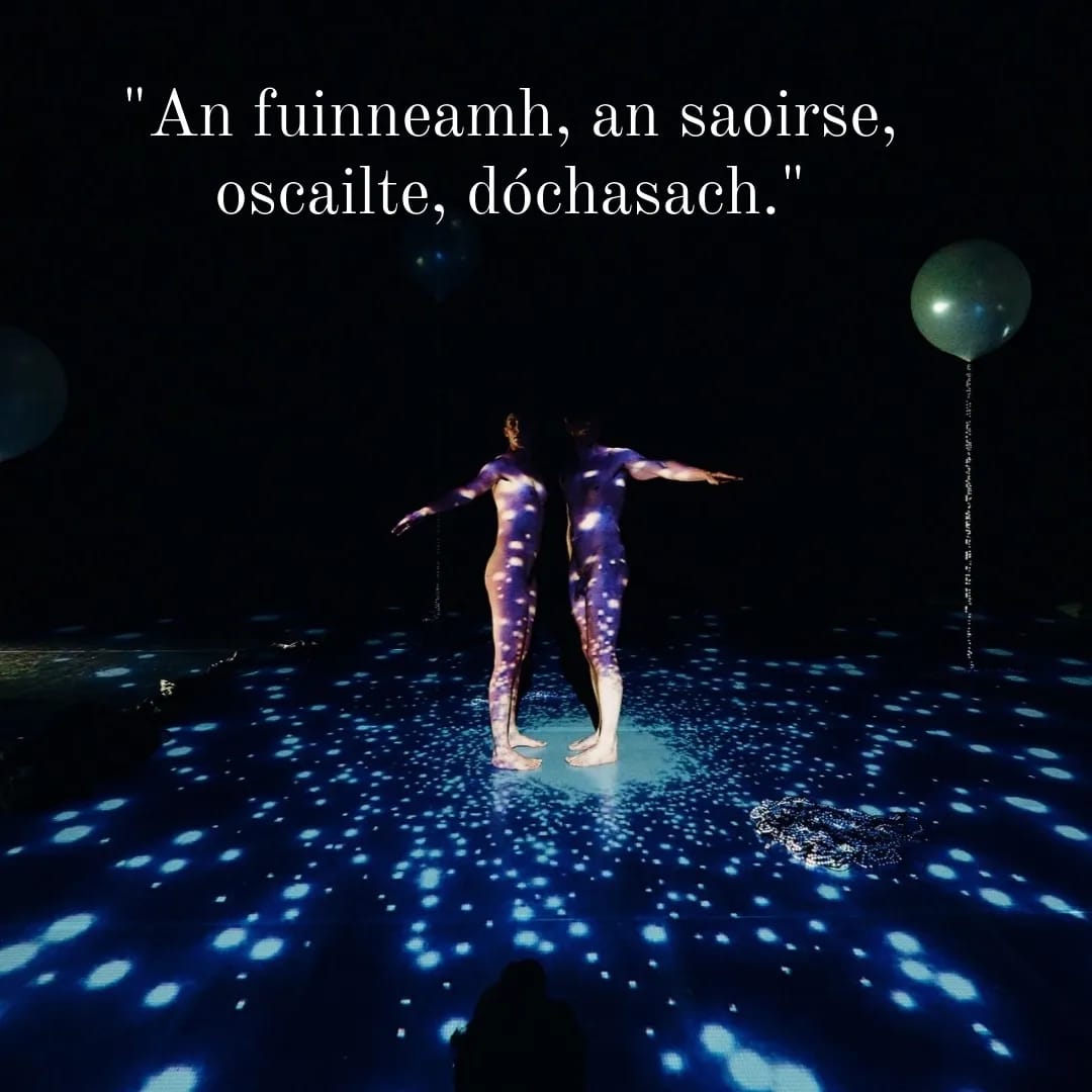 Two naked dancers stand with arms extended bathed in a projection of exploding particles. They are flanked by two white balloons with a string of pearls falling from them. Text above says: An fuinneamh, an Saoirse, oscailte, dóchasach"