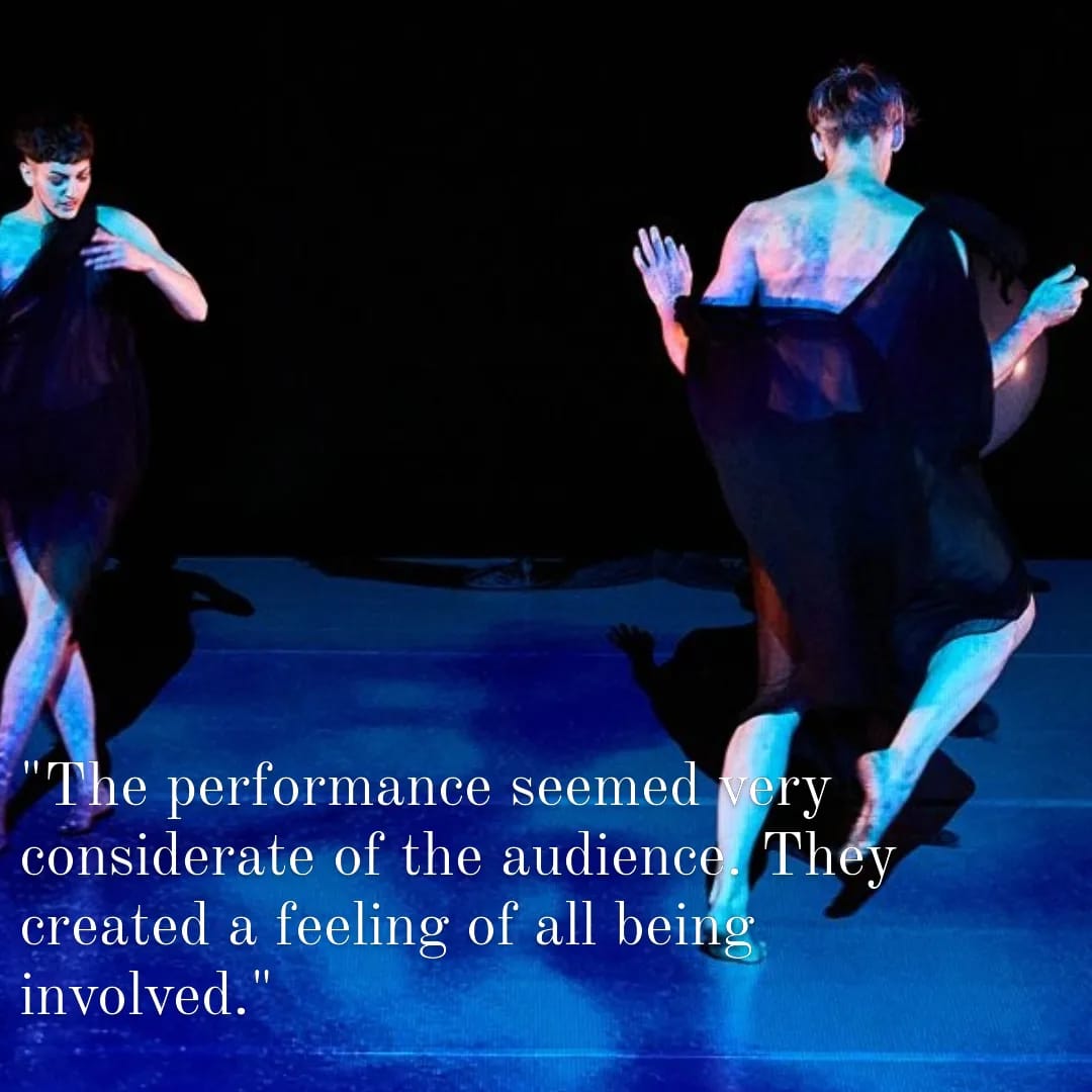 Two dancers in black toga like costumed dance upright, one back to the camera, the other facing the camera. They are bathed in a blue/purple projection. The text says: " The performance seemed very considerate of the audience. They created a feeling of all being involved."